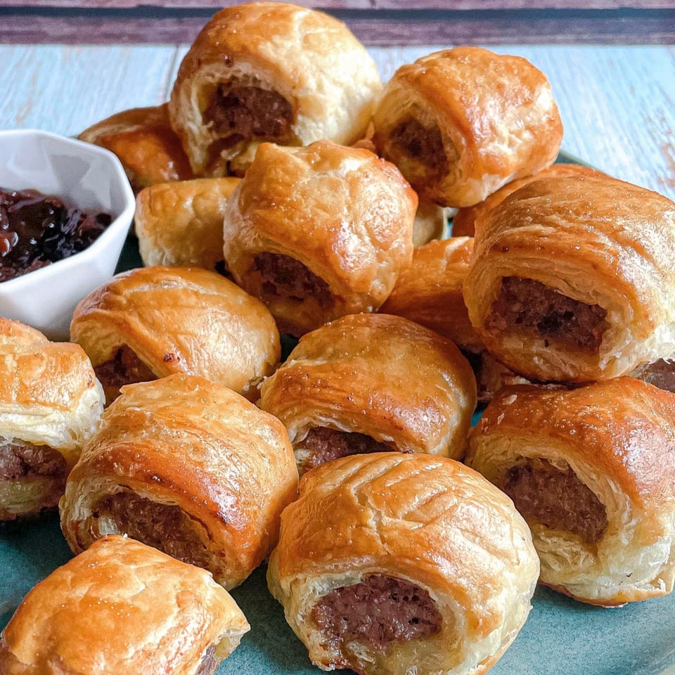 A platter of golden-brown sausage rolls with a side of dark chutney, ready to be served.