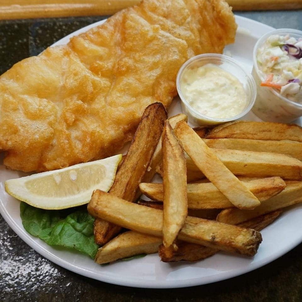Golden-brown fish and chips with tartar sauce, coleslaw, and a lemon wedge on a white plate.