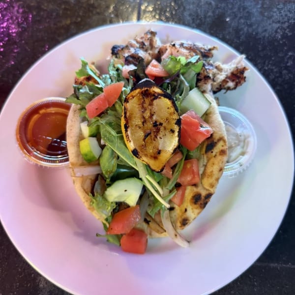 Grilled chicken pita with salad, charred lemon, and two dipping sauces on a white plate.